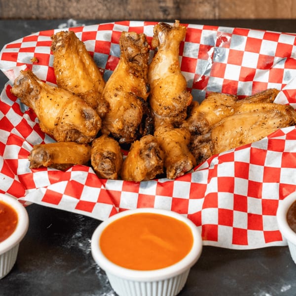 A basket of Uncle John's BBQ chicken wings with two dipping sauces on a dark surface.