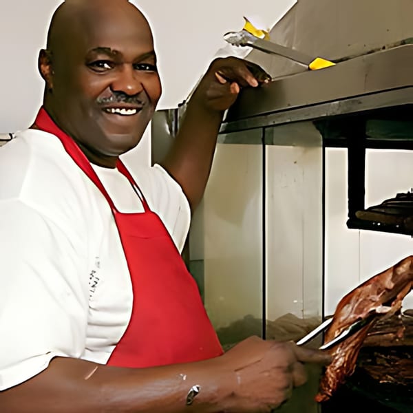 Smiling chef in a red apron tending to BBQ ribs on a grill at Uncle John's BBQ.