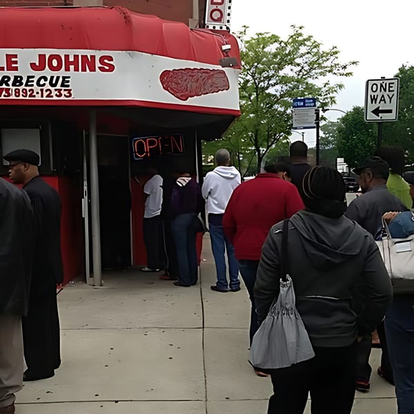 People queue outside Uncle John's BBQ, a red and white building with an 'OPEN' sign.