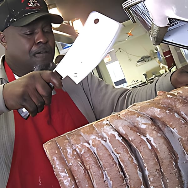 A man in a red apron and cap holds a cleaver over a large slab of ribs.