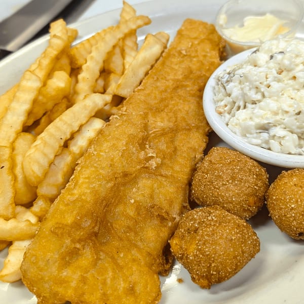 Fried fish, crinkle-cut fries, hush puppies, coleslaw, and tartar sauce on a white plate.