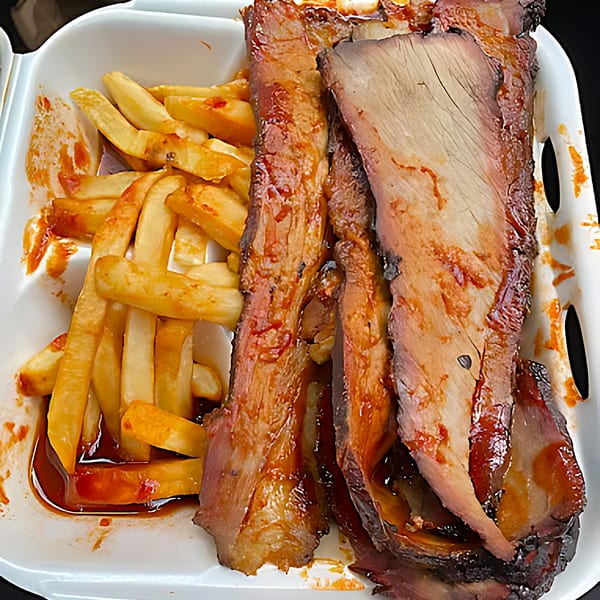 BBQ brisket and ribs with a side of french fries, drizzled with sauce in a takeout container.